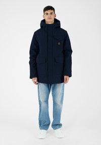 Giacca parka blu scuro con cappuccio, diverse tasche anteriori e un patch con logo sul petto. Indossata sopra jeans azzurri e sneaker bianche.