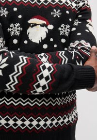 Sweater preto com um motivo de Pai Natal com óculos de sol, flocos de neve e padrões geométricos em vermelho e branco ao longo do peito e das mangas.
