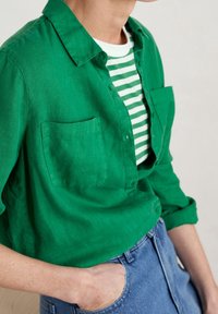 Groene linnen overhemd met knopen en twee borstzakken over een wit en groen gestreept T-shirt, gecombineerd met blauwe denim.