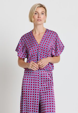 Femme aux cheveux blonds courts portant une combinaison à manches courtes aux motifs géométriques violets et bleus, debout les mains jointes.