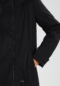 Veste noire à manches longues avec fermeture éclair frontale et poche poitrine zippée, main droite de la personne dans la poche latérale sur un fond clair uni.