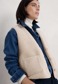 Bežna sherpa brezrokavnik z teksturiranim zaključkom, z sprednjim žepom in gumbnim zapiranjem, nosi se čez denimsko srajco in belo turtleneck majico.