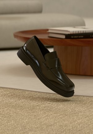 Mocassin en cuir noir flottant au-dessus d'un tapis beige texturé, avec une table basse ronde en bois et des livres en arrière-plan.