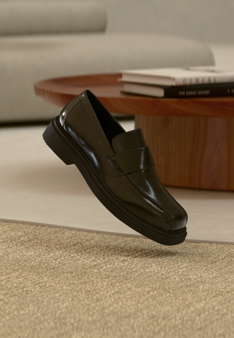 Mocassin en cuir noir flottant au-dessus d'un tapis beige texturé, avec une table basse ronde en bois et des livres en arrière-plan.