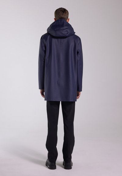 Man wearing a navy hooded raincoat, black pants, and black shoes, standing facing away against a plain light background.