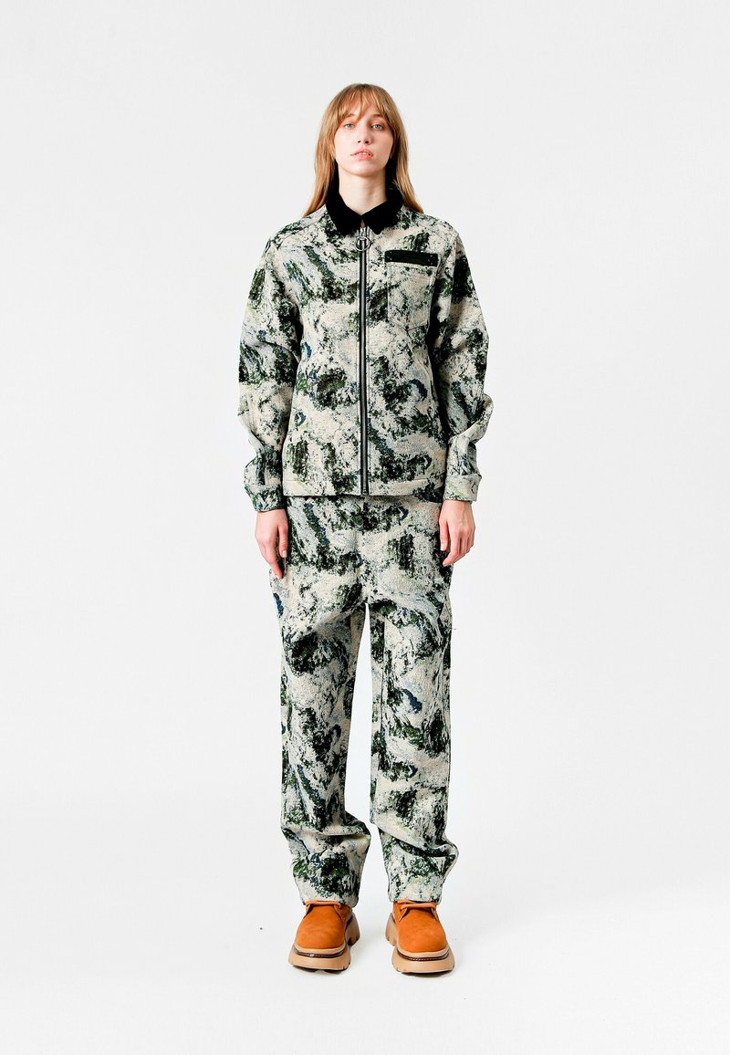 Conjunto texturizado que incluye una chaqueta con cuello y cremallera y pantalones a juego en un patrón abstracto verde y gris, combinados con zapatos marrones.