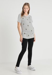 T-shirt riscada de maternidade em branco e preto, com abacaxis bordados, combinada com leggings pretas e ténis brancos.