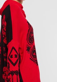 Rode en zwarte gebreide trui met een hoge kraag. Voorzien van decoratieve zwarte gedrukte ontwerpen en versieringen op de borst en mouwen.