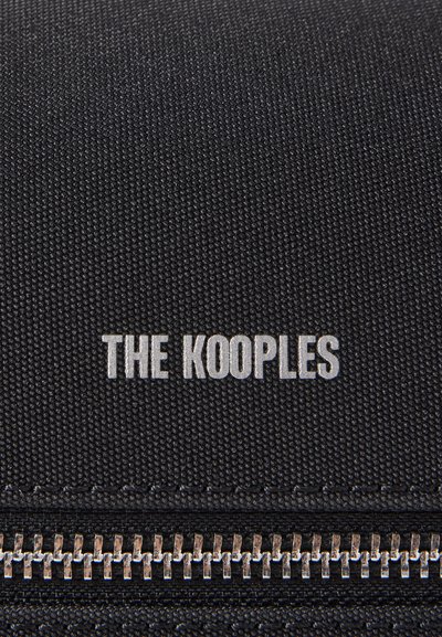 Melna auduma somiņa ar rāvējslēdzēju, teksturētu virsmu un sudraba "THE KOOPLES" logotipu. Apakšā ir metālisks rāvējslēdzēja aizdare.