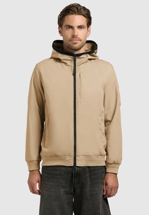 Man draagt een beige jas met rits en capuchon, met een zwarte binnenvoering en donkergrijze spijkerbroek, staand tegen een effen achtergrond.