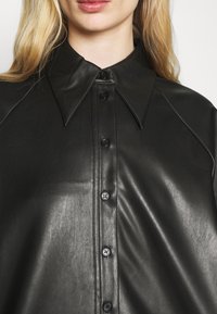 Camicia in pelle nera con un ampio colletto a punta. Presenta una chiusura frontale con bottoni neri e una texture liscia e lucida.