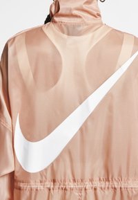 Ljust rosa vindjacka med en strukturerad yta, med en framträdande vit Nike-logga på baksidan och elastisk midja.