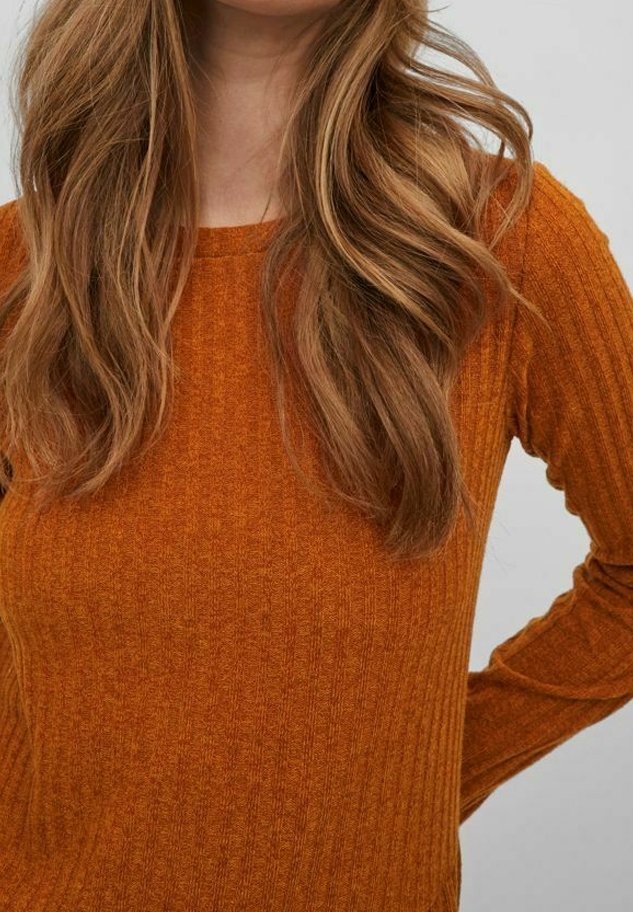 Femme aux longs cheveux ondulés châtain clair portant un pull à manches longues côtelé orange brûlée sur un fond clair uni.