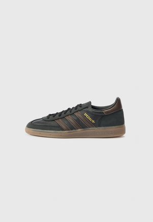 Zwarte Adidas Spezial sneaker met bruine strepen, een gumzool en gouden "Spezial" tekst aan de zijkant tegen een effen lichtgrijze achtergrond.