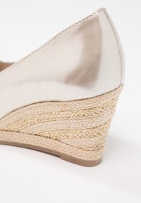 Chaussure à talon compensé en métal beige avec un talon en jute tissé présentant des accents dorés, une tige en cuir lisse et un design à bout pointu.
