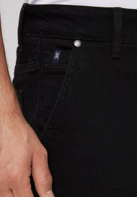 Schwarze Denim-Jeans im Slim-Fit-Schnitt, mit einem genähten Taschendesign, einem kleinen blauen Logo-Tag und einem Knopfverschluss am Bund.