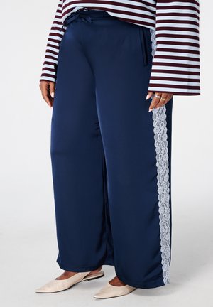 Pantalon large bleu marine avec une bande latérale en dentelle blanche, associé à un haut rayé à manches longues et des chaussures plates pointues beige.