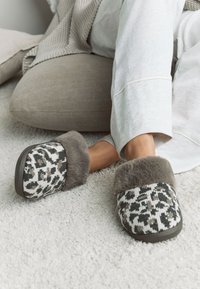 Chaussons tissés avec un imprimé animal noir et blanc, présentant une bordure en fourrure grise douce et une semelle en caoutchouc foncé, posés sur un tapis clair.