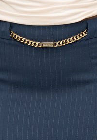 Σκούρα μπλε φούστα με διακριτικές ριγέ λεπτομέρειες, διαθέτει ζώνη με χρυσή αλυσίδα και ορθογώνιο μεταλλικό πόρπη. Απαλή υφή υφάσματος.