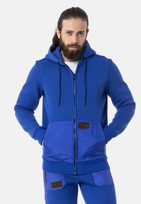 Blaues Zip-Hoodie mit Kängurutasche, strukturiertem Stoff und verstellbarem Kapuzenzug. Mit Lederpatch-Akzenten auf der Brust und an der Tasche.