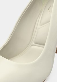 Chaussure blanche à talon haut avec un extérieur en cuir lisse. Caractérisée par un bout arrondi, une semelle intérieure texturée et un talon solide de couleur marron clair.