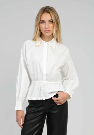 Chemise blanche à boutons avec col à volants et longues manches bouffantes, dotée d'une taille élastique et d'un ourlet festonné. Associée à un pantalon noir.