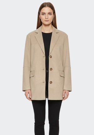 Donna che indossa un blazer beige monopetto con tre bottoni sopra una maglia nera e pantaloni neri, in piedi davanti a uno sfondo semplice.