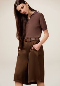 Brun kortærmet polo med en glitrende krave, parret med brede brune bukser med sidelommer og et matchende bælte.