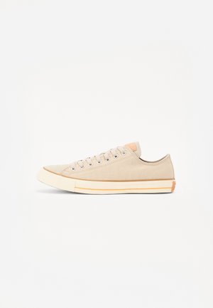 Beżowy niskoprofilowy sneaker z płótna z białą gumową podeszwą i sznurowadłami, subtelny pomarańczowy pasek na podeszwie i pomarańczowa łatka na pięcie, widok z boku.