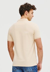 Beżowa koszulka polo z krótkim rękawem, żebrowanym kołnierzykiem i gładką fakturą, noszona z niebieskimi dżinsami. Skup się na widoku z tyłu i dopasowanym fasonie.