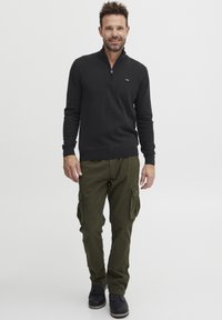 Suéter negro con cremallera en el cuello y mangas largas, combinado con pantalones cargo verde oliva que cuentan con bolsillos laterales. Botas negras completan el conjunto.