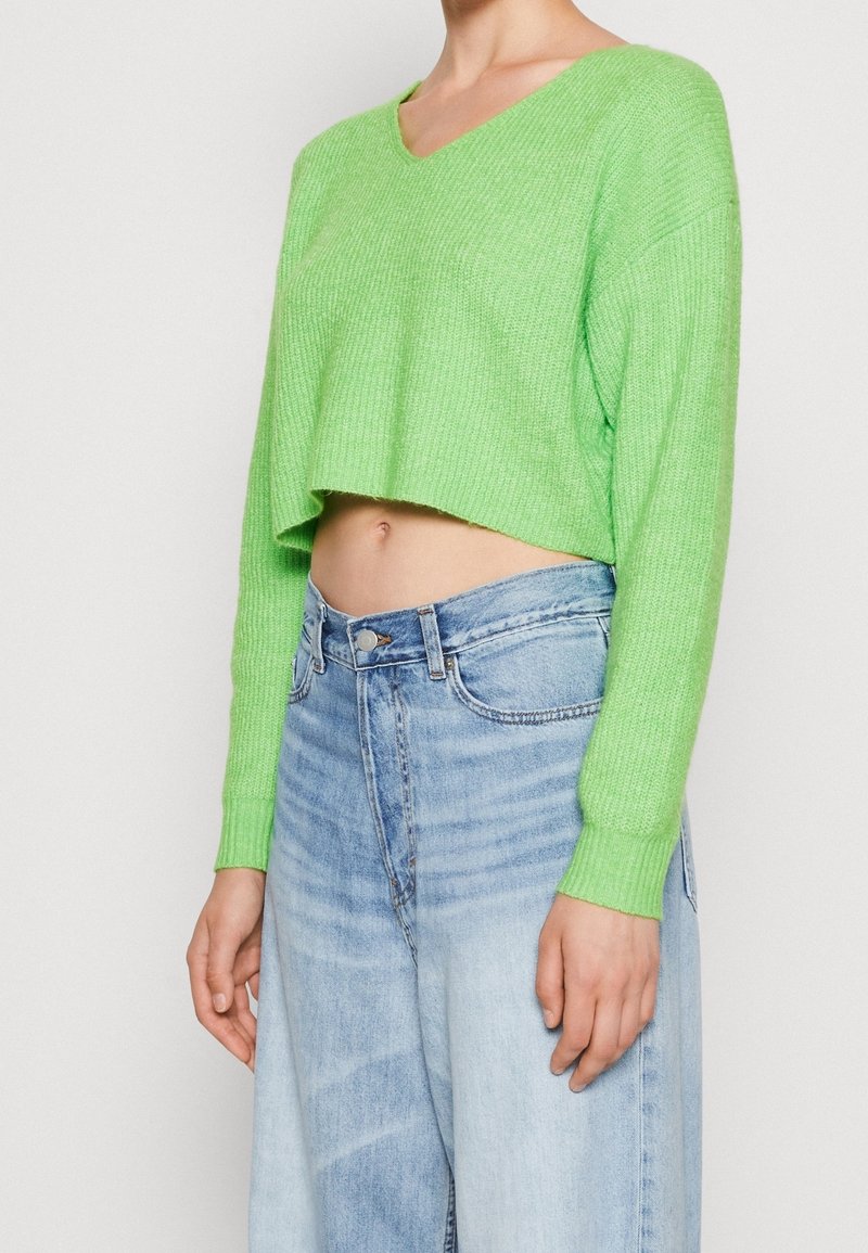 Femme portant un pull court en maille vert citron et un jean en denim bleu clair taille haute sur un fond uni.