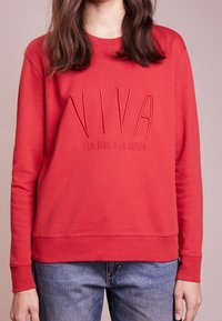 Žena v červené mikině s výšivkou "VIVA La Vida y La Fiesta" na přední straně, v kombinaci s modrými džíny, na světlém růžovém pozadí.