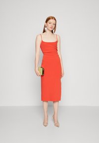 Red fitted midi dress with thin straps, smooth texture, and a tailored silhouette. Model holds a glittery gold clutch. Clear-heeled shoes.