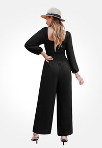 Czarny kombinezon z długimi rękawami i marszczonym stanikiem. Luźny, szeroki krój nogawek. Modelka nosi słomkowy kapelusz i srebrne sandały na obcasie.