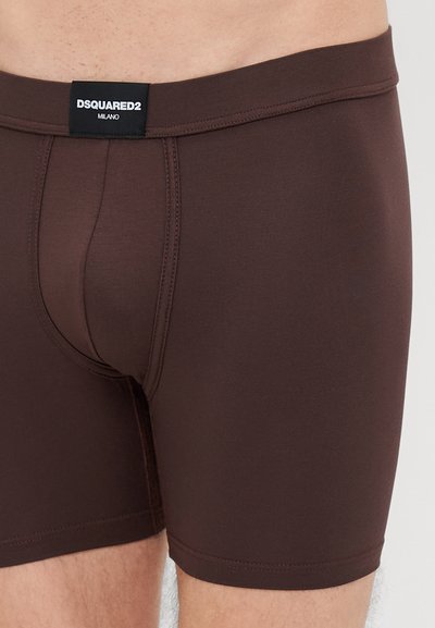 Καφέ μποξερ σορτς φτιαγμένα από ελαστικό ύφασμα. Διαθέτει μαύρη ζώνη με την επιγραφή "DSQUARED2 MILANO." Λείος υφή και εφαρμοστό σχέδιο.