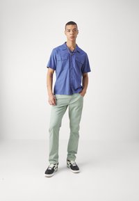 Camisa de botones de manga corta azul con dos bolsillos en el pecho, combinada con pantalones verdes claros y zapatillas negras con acentos estampados.