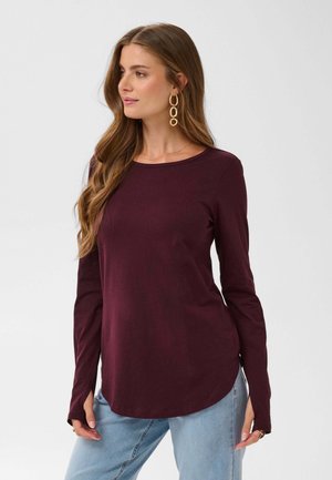 Femme aux longs cheveux ondulés portant un haut bordeaux à manches longues et un jean bleu clair, regardant sur le côté avec des boucles d’oreilles créoles dorées.