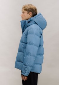 Blå puffjakke med hette, med quiltet design, glatt tekstur og frontlukking med glidelås. Modell vist fra siden.