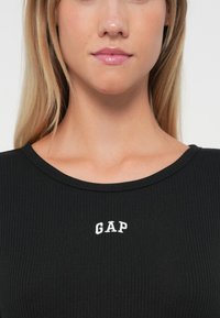 Crna rebrasta majica s okruglim izrezom i bijelim izvezenim "GAP" logotipom na prsima. Glatka tekstura i uski kroj.