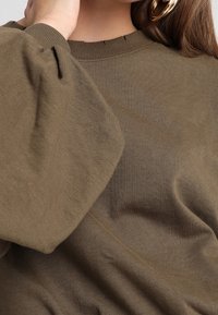 Suéter verde oliva de corte relajado, con cuello acanalado y mangas voluminosas. La tela parece suave y texturizada.