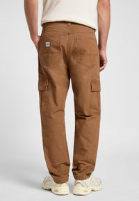 Pantalones cargo marrones de algodón con dos bolsillos laterales, dos bolsillos traseros y un parche con el logo en la cinturilla trasera. Combinados con zapatillas ligeras.