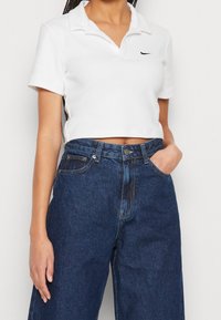 Kvinna iklädd en vit kortärmad polo och högmidjade, lösa mörkblå jeans, med ena handen i fickan mot en enfärgad bakgrund.