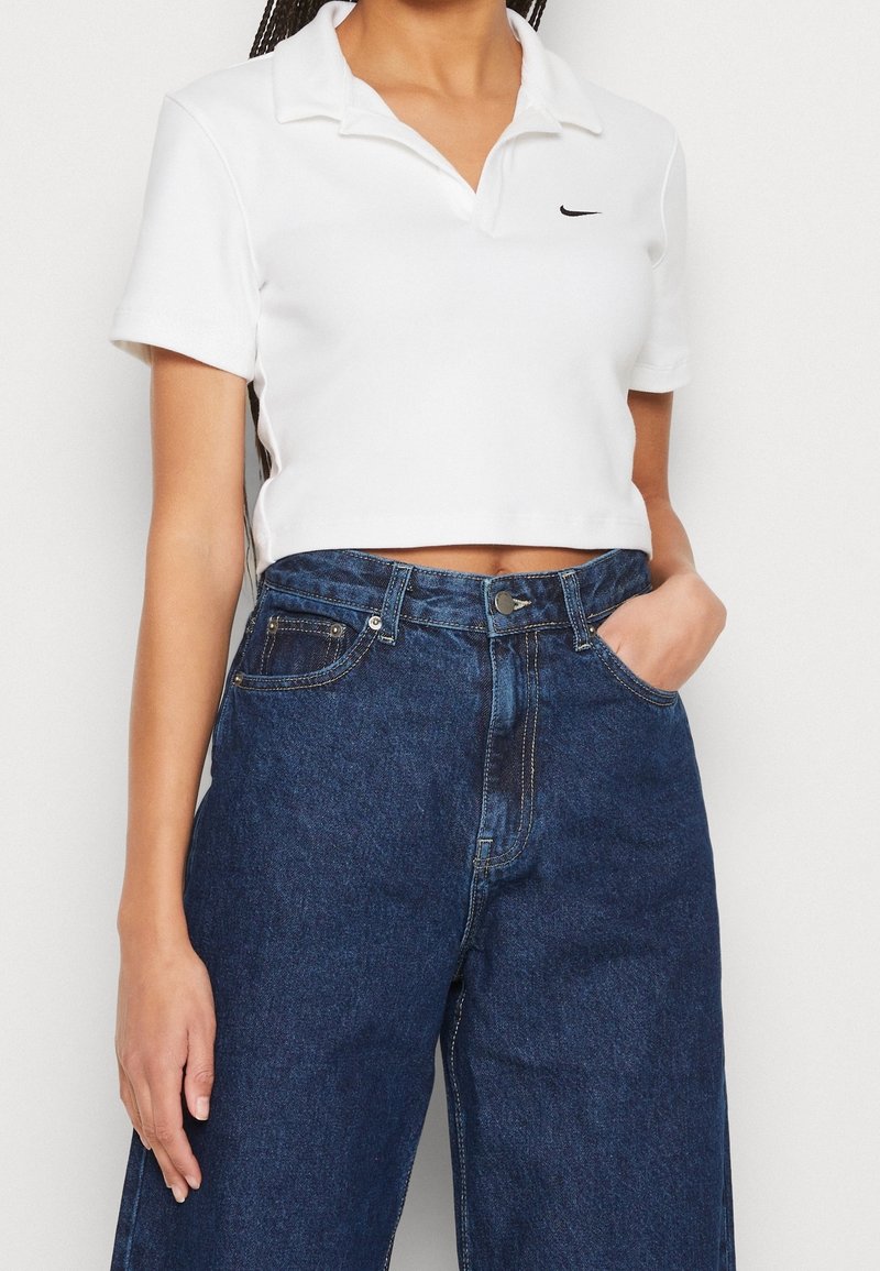 Kvinna iklädd en vit kortärmad polo och högmidjade, lösa mörkblå jeans, med ena handen i fickan mot en enfärgad bakgrund.