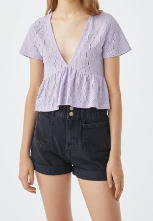 Femme portant un haut péplum texturé lavande à manches courtes et encolure en V, associé à un short en denim noir taille haute avec revers roulés et poches avant.