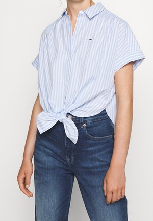 Blauw-wit gestreept overhemd met korte mouwen, geknoopt in de taille, gedragen met donkerblauwe spijkerbroek. Voorzien van een kraag en geborduurd logo.