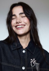 Una giovane donna con lunghi capelli scuri sorride a occhi chiusi, indossando una giacca di jeans scura decorata con una spilla a forma di fiore gioiello.