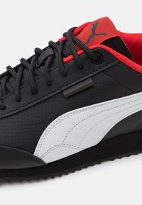 Chaussure de sport noire avec une finition en cuir lisse, accents rouges au talon, logo blanc sur le côté, et semelle extérieure en caoutchouc noir texturée.