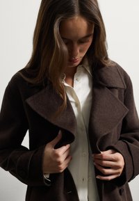 Bruine wollen blendjas met grote kleppen, getailleerde pasvorm, knopenaccenten en een zachte textuur, gedragen over een wit shirt.