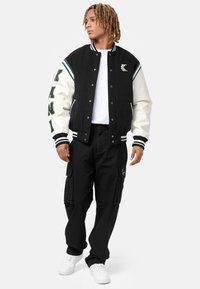 Jachetă varsity neagră și crem cu accente verzi, inscripții mari și manșete cu dungi, asortată cu pantaloni cargo negri și adidași albi.